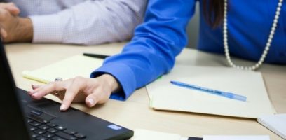 Two people sit at a desk with papers, folders, and a pen. One person, wearing a blue shirt and a pearl necklace, is using a laptop, while the other persons arm is partially visible in the background.