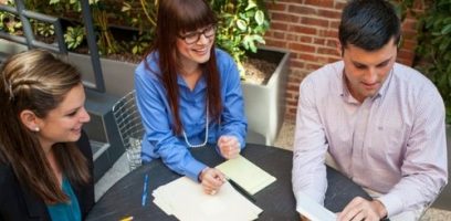 Three people sit around a round table outdoors with papers, pens, and notebooks, having a discussion. Two women and one man appear engaged and smiling amid greenery and a brick wall background.
