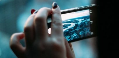 A close-up of a person holding a smartphone horizontally to take a photo of a cityscape with water and buildings in the background. The person’s fingernails are painted dark and light colors.