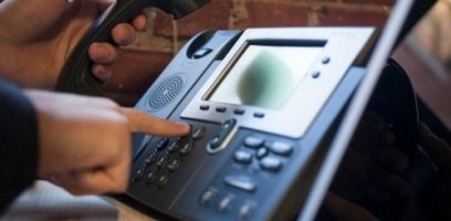 customer_service_blog_8 A person holds a phone receiver and presses buttons on an office desk phone with a digital display, set against a brick wall background, representing the professional touch provided by Abby Connect.