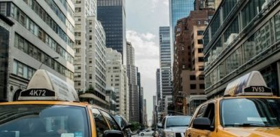 Yellow taxis drive down a busy city street lined with tall skyscrapers and office buildings. The sky is partly cloudy, and other vehicles are visible between the taxis.