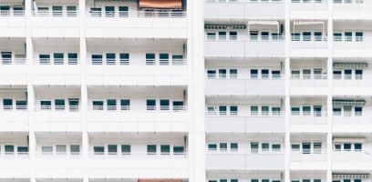 A white apartment building facade with rows of identical windows and balconies, viewed straight on. Some balconies have awnings, and the overall design is very geometric and repetitive.