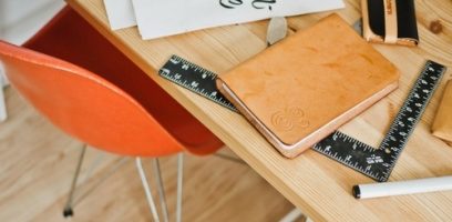 A wooden desk with a tan notebook, two black rulers, a white marker, and paper on top. An orange chair is positioned next to the desk on a light wood floor.