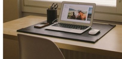 A laptop displaying a photo of a cow sits on a desk next to a smartphone, a cup filled with pens and scissors, and a wireless mouse; a chair is pushed in and a window is in the background.