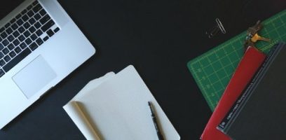 A laptop, an open notebook with a pen, a green cutting mat, paper clips, keys, and two closed folders are arranged neatly on a dark desk.