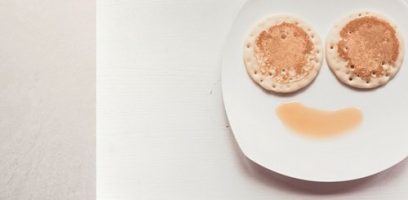 A white plate with two pancakes arranged as eyes and a curved line of syrup below them, creating a smiling face on a light background.