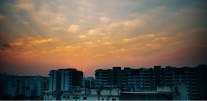 A cityscape silhouette of tall buildings under a colorful sunset sky, with rays of sunlight shining through scattered clouds.