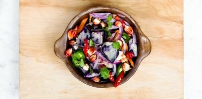 A brown bowl filled with a colorful salad featuring red onions, cherry tomatoes, leafy greens, sliced chili peppers, chickpeas, and a creamy dressing, placed on a wooden surface.
