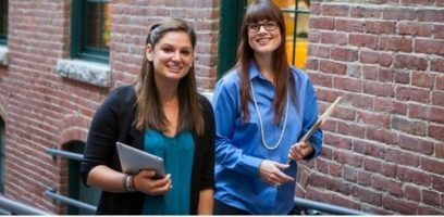 customer_service_blog_98 Two women smiling, walking outdoors next to a brick wall. One holds a tablet and the other carries a folder and papers. Both appear to be in a casual, professional setting.