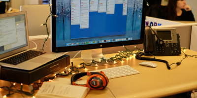 A decorated office desk with a computer monitor, keyboard, phone, laptop, headphones, and notebook. Holiday string lights are draped around the desk. In the background, a person sits at another workstation.