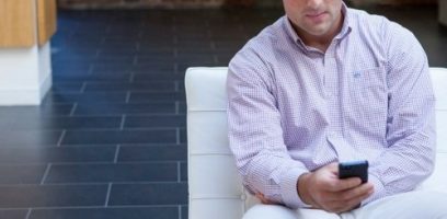 email_newsletter_subimages_500x245_4 A man in a light purple checkered shirt sits on a white sofa, looking at a smartphone in his hand. The background features dark tiled flooring and part of a light-colored wall.