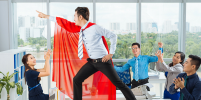 A man wearing a red superhero cape strikes a heroic pose on a desk in an office, while four coworkers cheer, clap, and smile at him. Large windows show a cityscape in the background.