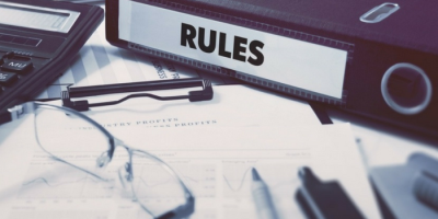 A binder labeled RULES sits on a desk alongside papers, a calculator, eyeglasses, and a pen, suggesting an office or business setting focused on regulations or compliance.