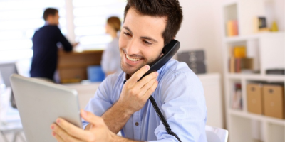 A smiling man sits at a desk, holding a landline phone to his ear while looking at a tablet. He appears engaged and happy. In the background, two people converse in a bright, modern office setting.
