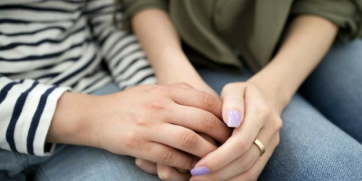 Close-up of two people sitting side by side, gently holding hands. One is wearing a striped shirt, the other an olive top and a ring, with light purple nail polish. Both are dressed in jeans.