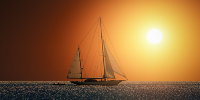 A sailboat glides across a calm sea at sunset, with the bright sun low on the horizon and the sky glowing in warm orange tones. The water shimmers with reflected light.