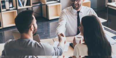 A smiling man in a shirt and tie shakes hands with another man across a desk, while a woman holding a coffee cup sits beside them. In this modern office setting, their teamwork clearly outshines the competition.