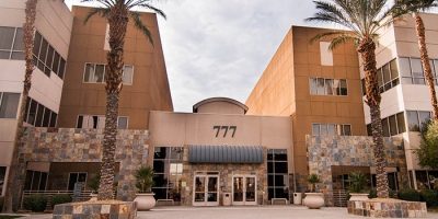 The entrance of a modern office building, home to Abby Connect, features large windows, palm trees, a stone facade, and the number 777 above the doors. The building showcases a tan and brown exterior.