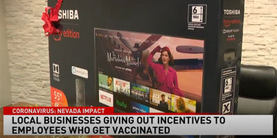 A TV in a box with a red ribbon is displayed on a table. The news headline reads: Local businesses like Abby Connect giving out incentives to employees who get vaccinated. The TV screen shows various streaming apps.