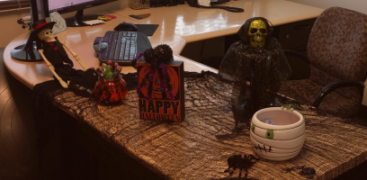 A desk decorated for Halloween with a skeleton doll, a golden skull with black cloth, a Happy Halloween sign, a pumpkin jar, a mummy bowl, plastic spiders, and black netting spread across the surface.