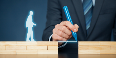 A person in a suit draws a blue line between two stacks of wooden blocks, with a glowing silhouette of a human figure walking toward the gap.