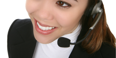 A smiling woman wearing a headset and business attire, suggesting she is working in customer service or a call center. The photo is taken from an angle above her left shoulder.