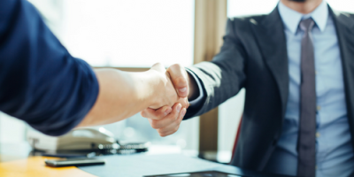 Two people are shaking hands across a desk in an office setting, one wearing a suit and the other in casual clothing.