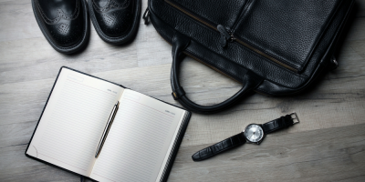 Flat lay of black leather shoes, a black leather briefcase, an open notebook with a pen, and a black leather wristwatch on a wooden floor.
