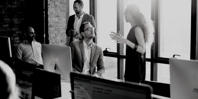 Black and white photo of four people in an office, having a discussion near large windows. One woman is standing and gesturing, while three men sit and listen, with computers and reports visible on desks.
