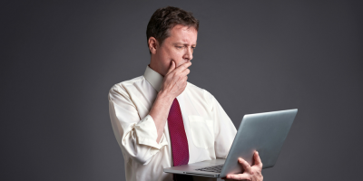 A man in a white shirt and red tie holds a laptop in one hand while looking at the screen with a thoughtful expression, his other hand touching his chin, standing against a gray background.