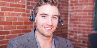 A smiling man wearing a headset sits in front of an exposed brick wall, dressed in a grey blazer and light shirt, appearing to work in a call center or customer service environment.