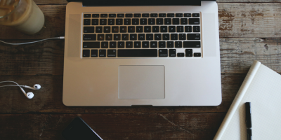 A silver laptop on a rustic wooden table, surrounded by white earphones, a smartphone, a notebook, and a glass of iced coffee—perfect for staying productive while managing your real estate business.