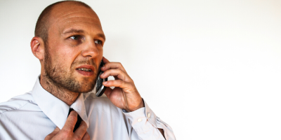 A man in a dress shirt adjusts his tie with one hand while talking on a mobile phone, looking slightly concerned. The background is plain white.