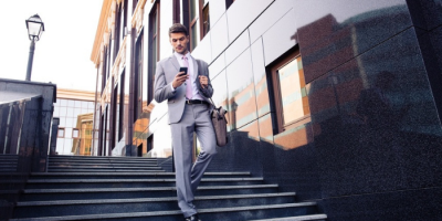 A man in a gray suit walks down outdoor steps near a modern office building, looking at his phone and carrying a brown bag.