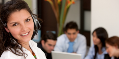 A woman wearing a headset smiles at the camera in the foreground, while four people sit together at a table in the background, collaborating with a laptop.