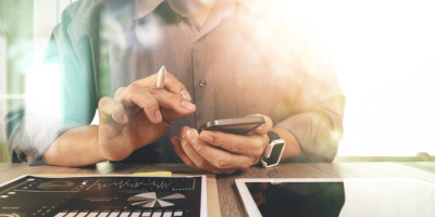 A person in a button-up shirt uses a smartphone while working at a desk with business charts, a tablet, and a smartwatch, with sunlight streaming in from the right.