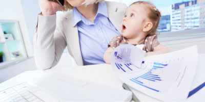A woman in business attire holds a baby in her lap while talking on the phone at a desk with charts and a laptop, illustrating work-life balance. The baby looks upward with an open mouth expression.